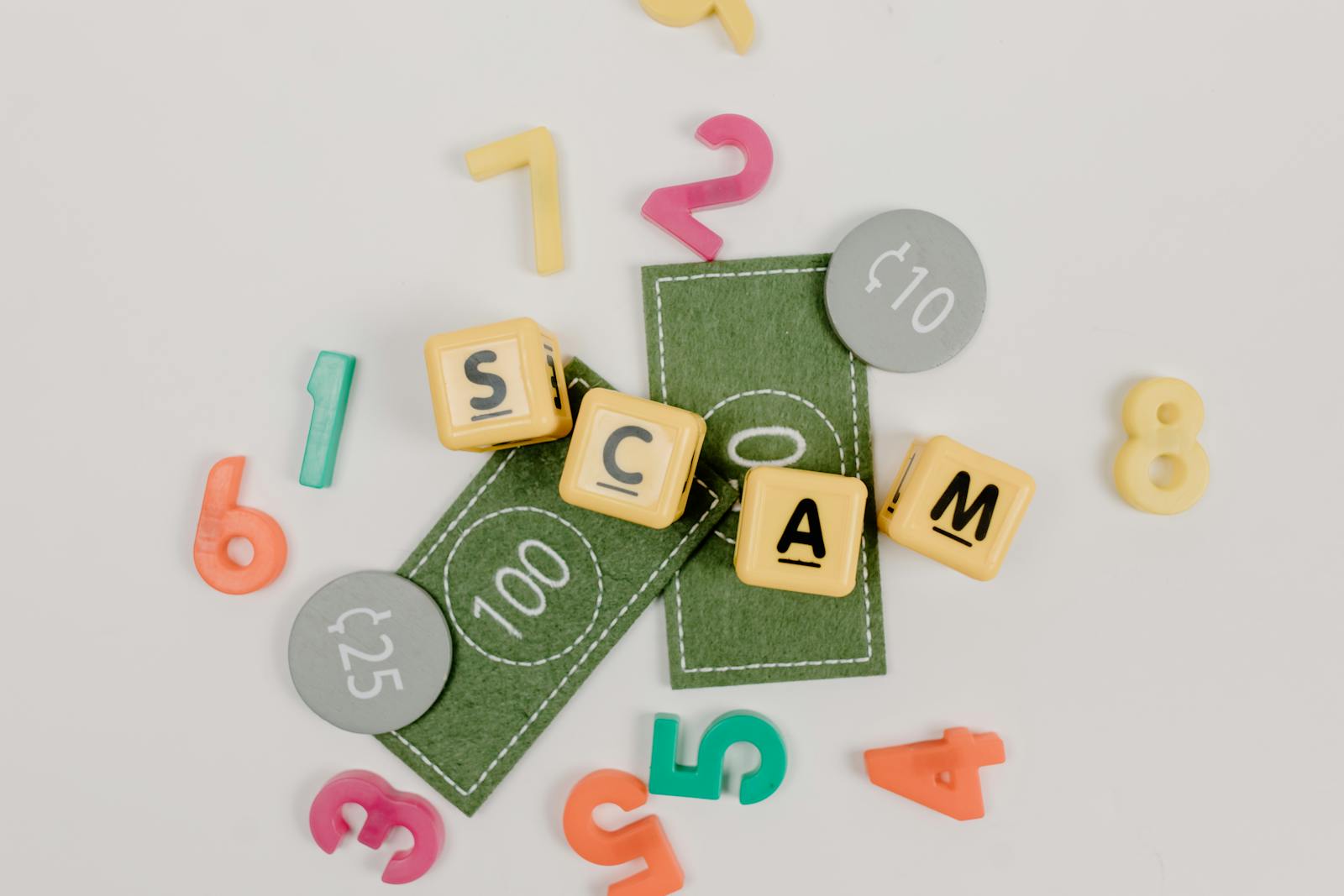 Scrabble tiles spelling 'scam' with play money and numbers on a white background.